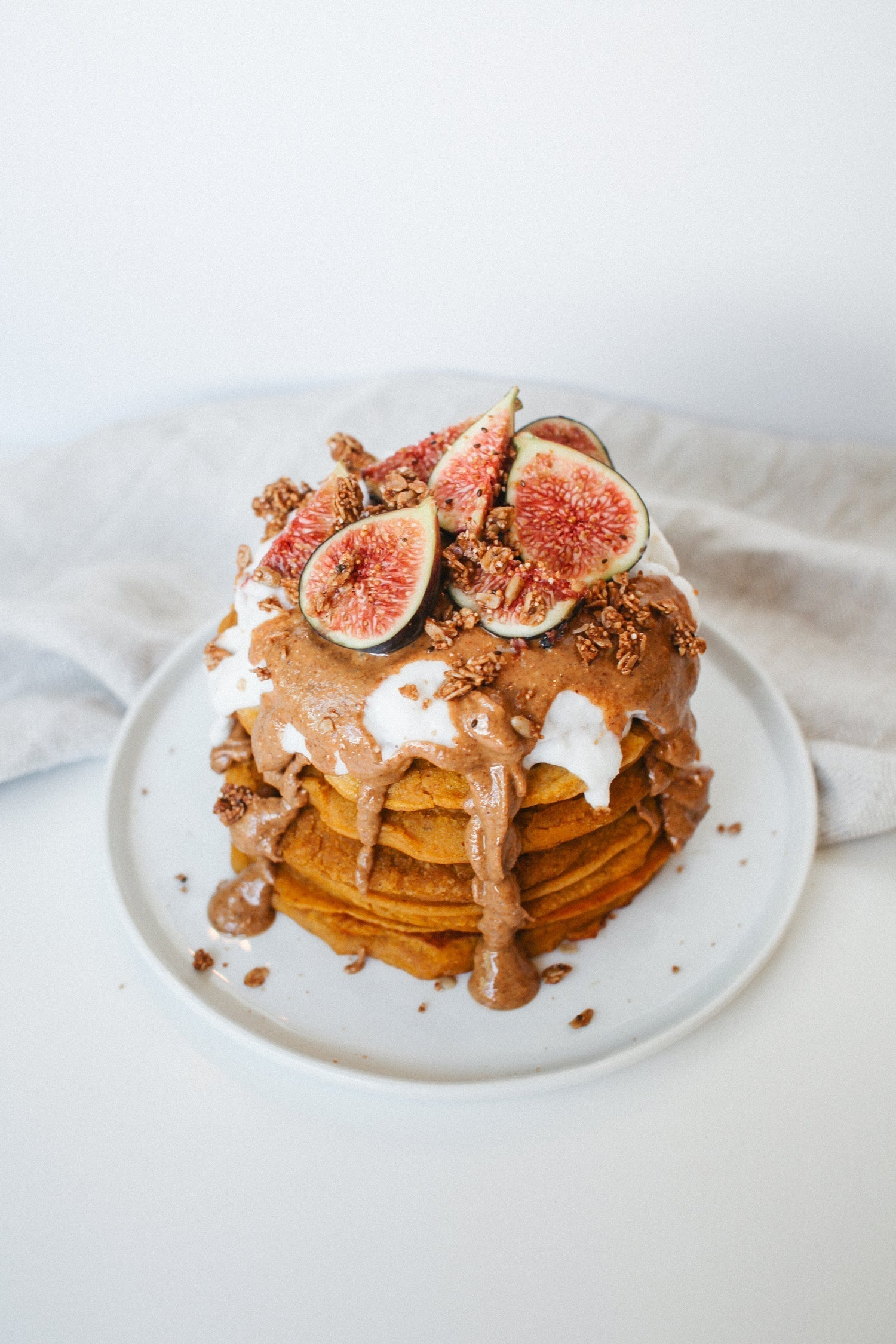 top view of a white round plate with a pile of pumpkin pancakes with whip cream and chocolate ganache topped with choco bits on top of a white table with white cloth on the side