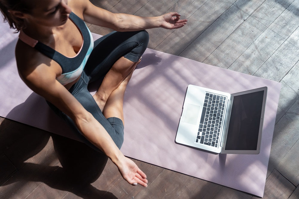 Woman meditating in front of her laptop while sitting on a pink yoga mat