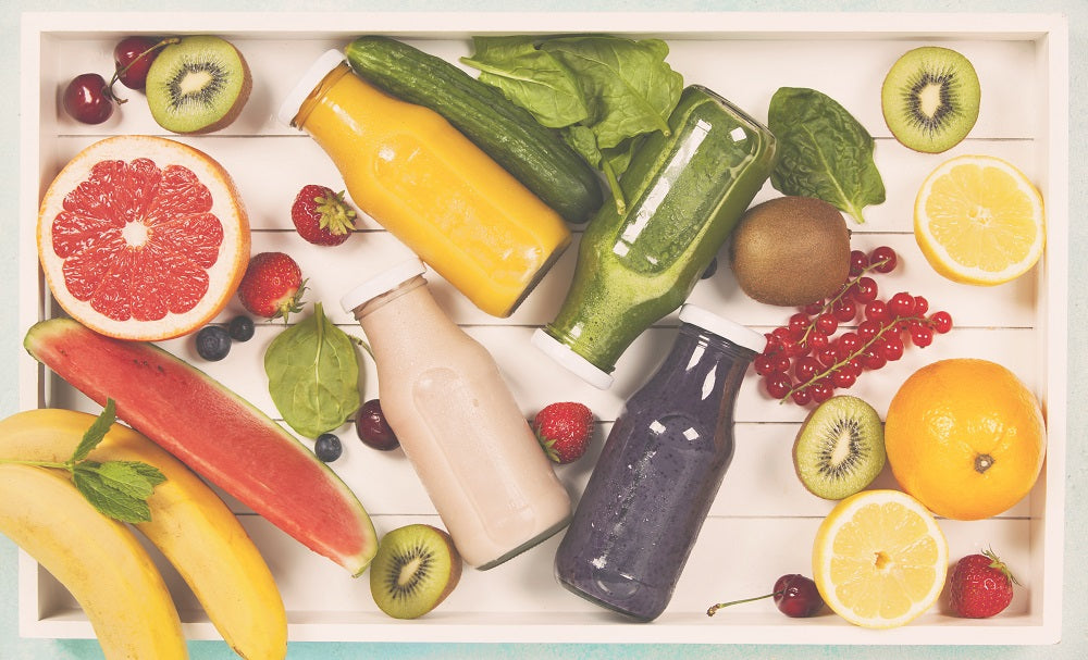 A top view shot of 4 jars of different smoothies surrounded by half a grape fruit, slices of kiwi, slices of orange, slices of lemon, 2 bananas, a slice of watermelon, strawberries, and grapes.