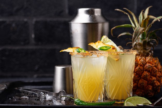 two glasses of pineapple chia margarita with cut pineapple and a sliced of chili on top surrounded by cut lemon, chili, pineapple, a stainless pitcher, stainless glass, and a stainless tong