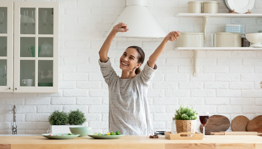 Image of a happy woman in her kitchen, waving her hands in the air.
