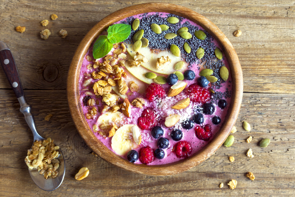 a bowl of vegan smoothie sprinkled with squash seeds, assorted nuts, mulberry, blueberry, banana, and a spoon with rice flakes on the side of the bowl and scattered nuts on top of the table