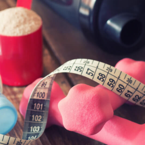 two small pink dumbbell and a tape measure on the top of it, with a red cup full of white powder of Pea protein, and large blue tumbler on the side on top of a wooden table