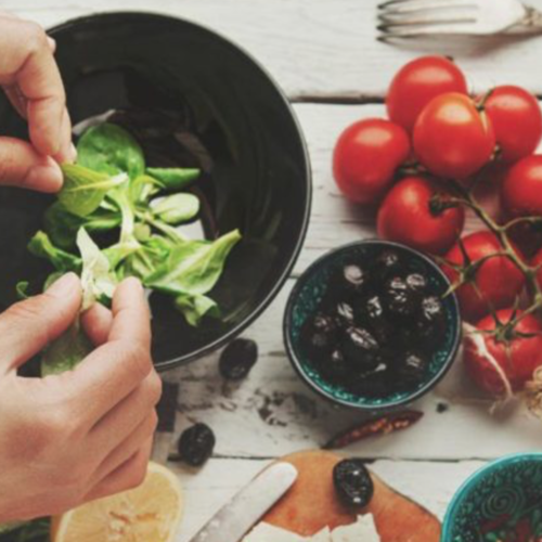 Top view shot of a woman's hands putting some more green vegetables in a bowl of salad. Beside the bowl of salad is a smaller bowl of dried olives and a bunch of tomatoes still attached to its stem.